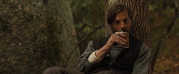 Movie still from “Redeeming Love” (2022), directed by D.J. Caruso – A man sitting on the ground drinking a cup of coffee; Medium shot, Over the shoulder angle