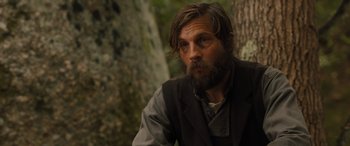 Movie still from “Redeeming Love” (2022), directed by D.J. Caruso – A man with long hair and a goatee sitting in front of a tree; Close Up shot, Over the shoulder angle