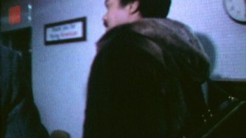Movie still from “Reggie” (2023), directed by Alex Stapleton – A man wearing a fur coat standing next to a wall; Medium shot, Over the shoulder angle