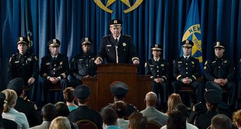 Movie still from “Bad Lieutenant: Port of Call New Orleans” (2009), directed by Werner Herzog – A group of people sitting in front of a man in uniform; Wide shot, Over the shoulder angle