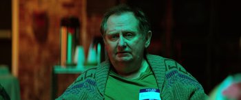 Movie still from “Renfield” (2023), directed by Chris McKay – An older man wearing a green shirt and a green sweater; Close Up shot, Over the shoulder angle