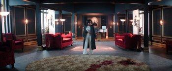 Movie still from “Renfield” (2023), directed by Chris McKay – A woman standing in a living room wearing a long coat; Wide shot, High angle