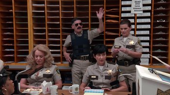 Movie still from “Reno 911!: The Hunt for QAnon” (2021), directed by Robert Ben Garant – A group of police officers sitting at a table; Wide shot, Over the shoulder angle