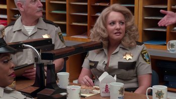 Movie still from “Reno 911!: The Hunt for QAnon” (2021), directed by Robert Ben Garant – A woman sitting at a table with a box of food in front of her; Medium shot, Over the shoulder angle