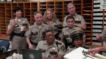 Movie still from “Reno 911!: The Hunt for QAnon” (2021), directed by Robert Ben Garant – A group of police officers sitting and standing in front of a laptop; Medium shot, High angle
