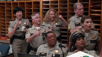 Movie still from “Reno 911!: The Hunt for QAnon” (2021), directed by Robert Ben Garant – A group of people in police uniforms singing together; Medium shot, High angle