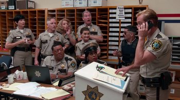 Movie still from “Reno 911!: The Hunt for QAnon” (2021), directed by Robert Ben Garant – A group of police officers gathered around a box; Medium shot, High angle