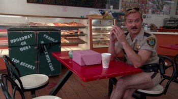 Movie still from “Reno 911!: The Hunt for QAnon” (2021), directed by Robert Ben Garant – A man sitting at a table in front of a box of donuts; Wide shot, Over the shoulder angle