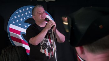 Movie still from “Reno 911!: The Hunt for QAnon” (2021), directed by Robert Ben Garant – A man holding a microphone while standing in front of an audience; Medium shot, Low angle