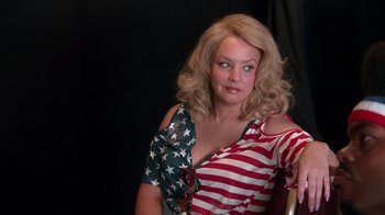 Movie still from “Reno 911!: The Hunt for QAnon” (2021), directed by Robert Ben Garant – A woman with blonde curly hair wearing an american flag shirt; Medium shot, Over the shoulder angle