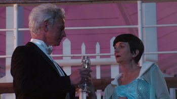 Movie still from “Reno 911!: The Hunt for QAnon” (2021), directed by Robert Ben Garant – An older man and a younger woman holding champagne flutes in front of a fence; Medium shot, Low angle