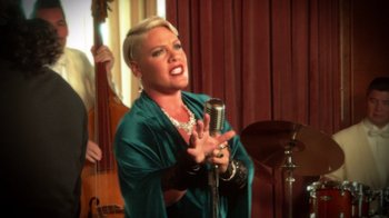 Movie still from “Reno 911!: The Hunt for QAnon” (2021), directed by Robert Ben Garant – A woman singing into a microphone in front of a microphone stand; Medium shot, Over the shoulder angle