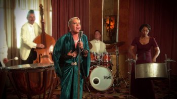 Movie still from “Reno 911!: The Hunt for QAnon” (2021), directed by Robert Ben Garant – A woman singing into a microphone in front of a drum set; Wide shot, Low angle