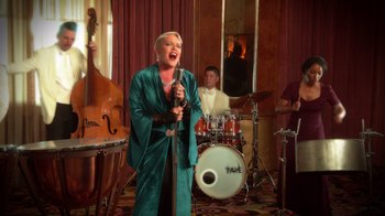 Movie still from “Reno 911!: The Hunt for QAnon” (2021), directed by Robert Ben Garant – A woman singing into a microphone in front of a drum set; Wide shot, Low angle