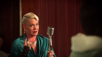 Movie still from “Reno 911!: The Hunt for QAnon” (2021), directed by Robert Ben Garant – A woman holding a microphone in front of a microphone stand; Close Up shot, Over the shoulder angle