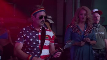Movie still from “Reno 911!: The Hunt for QAnon” (2021), directed by Robert Ben Garant – A man in a patriotic shirt playing a guitar; Medium shot, Over the shoulder angle