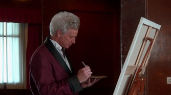 Movie still from “Reno 911!: The Hunt for QAnon” (2021), directed by Robert Ben Garant – A man in a suit writing on a piece of paper in front of an easel; Medium shot, Low angle