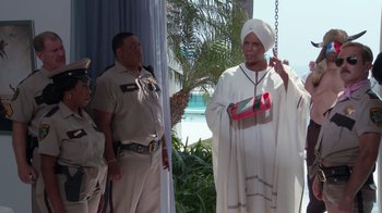 Movie still from “Reno 911!: The Hunt for QAnon” (2021), directed by Robert Ben Garant – A man in a white robe holding onto a pink purse; Medium shot, Over the shoulder angle