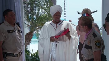 Movie still from “Reno 911!: The Hunt for QAnon” (2021), directed by Robert Ben Garant – A woman in a white robe holding a red box; Medium shot, Over the shoulder angle