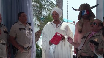 Movie still from “Reno 911!: The Hunt for QAnon” (2021), directed by Robert Ben Garant – A woman in a white robe holding a red bag; Medium shot, Over the shoulder angle