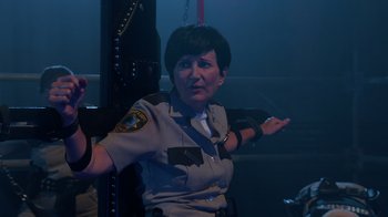Movie still from “Reno 911!: The Hunt for QAnon” (2021), directed by Robert Ben Garant – A woman in a police uniform standing in front of a metal pole; Medium shot, Over the shoulder angle