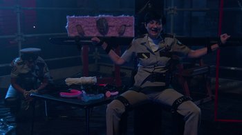 Movie still from “Reno 911!: The Hunt for QAnon” (2021), directed by Robert Ben Garant – A woman sitting in a chair with a knife in her hand; Medium shot, High angle