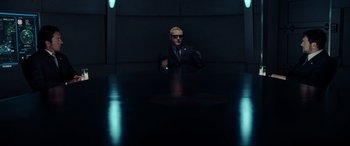 Movie still from “Resident Evil: Extinction” (2007), directed by Russell Mulcahy – A man in a suit and sunglasses sitting at a table in a dark room; Wide shot, Over the shoulder angle