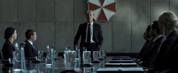 Movie still from “Resident Evil: The Final Chapter” (2016), directed by Paul W.S. Anderson – A man in a suit and tie standing in front of a table; Medium shot, Over the shoulder angle