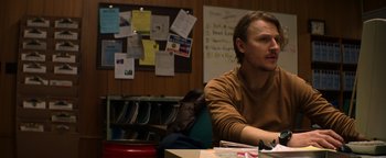 Movie still from “Resident Evil: Welcome to Raccoon City” (2021), directed by Johannes Roberts – A man sitting at a table in front of papers on the wall; Medium shot, Over the shoulder angle