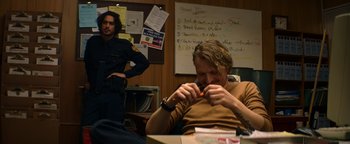 Movie still from “Resident Evil: Welcome to Raccoon City” (2021), directed by Johannes Roberts – A man sitting at a table with a man standing next to him; Medium shot, Over the shoulder angle