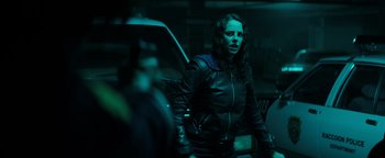 Movie still from “Resident Evil: Welcome to Raccoon City” (2021), directed by Johannes Roberts – A woman standing next to a car in a dark room; Medium shot, Over the shoulder angle