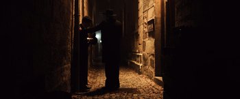 Movie still from “Resistance” (2020), directed by Jonathan Jakubowicz – A man in a black suit and a woman in a black hat in an alleyway at night; Wide shot, Over the shoulder angle
