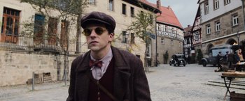 Movie still from “Resistance” (2020), directed by Jonathan Jakubowicz – A man in a hat and bow tie walking down a street; Medium shot, Over the shoulder angle