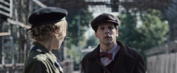Movie still from “Resistance” (2020), directed by Jonathan Jakubowicz – A man in a hat and bow tie standing next to a woman; Close Up shot, Over the shoulder angle