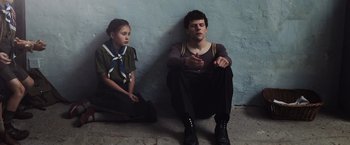 Movie still from “Resistance” (2020), directed by Jonathan Jakubowicz – A man sitting next to a young girl in front of a blue wall; Medium shot, Low angle
