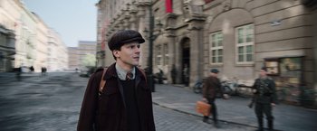 Movie still from “Resistance” (2020), directed by Jonathan Jakubowicz – A man in a hat and a brown jacket walking down a street; Medium shot, Over the shoulder angle
