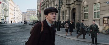 Movie still from “Resistance” (2020), directed by Jonathan Jakubowicz – A man wearing a hat walking down a street; Medium shot, Over the shoulder angle