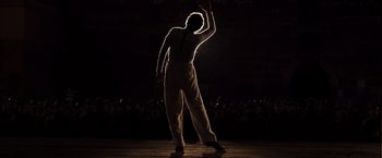 Movie still from “Resistance” (2020), directed by Jonathan Jakubowicz – A man in a striped shirt and white pants is on a stage waving; Wide shot, Low angle