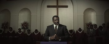 Movie still from “Respect” (2021), directed by Liesl Tommy – A man in a suit and tie in front of a cross; Medium shot, Low angle
