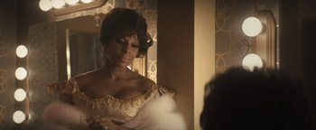 Movie still from “Respect” (2021), directed by Liesl Tommy – A black woman in a gold dress with a white feather duster; Close Up shot, Over the shoulder angle