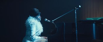Movie still from “Respect” (2021), directed by Liesl Tommy – A person sitting at a microphone next to a grand piano; Medium shot, Over the shoulder angle