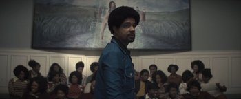 Movie still from “Respect” (2021), directed by Liesl Tommy – A group of people sitting in a room with a man standing in front of a painting; Medium shot, Low angle