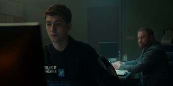 Movie still from “Restless” (2022), directed by Régis Blondeau – A man in a police uniform sitting in front of a computer; Medium shot, Over the shoulder angle
