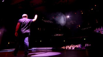 Movie still from “Ricky Gervais: SuperNature” (2022), directed by John L. Spencer – A man standing in front of an audience on a stage; Wide shot, High angle