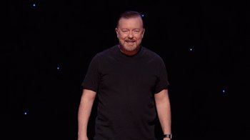 Movie still from “Ricky Gervais: SuperNature” (2022), directed by John L. Spencer – A man in a black t - shirt standing in front of a black background; Medium shot, Low angle