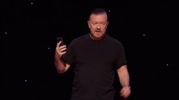 Movie still from “Ricky Gervais: SuperNature” (2022), directed by John L. Spencer – A man in black shirt holding a cell phone; Medium shot, High angle