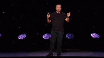 Movie still from “Ricky Gervais: SuperNature” (2022), directed by John L. Spencer – A man standing on a stage holding a cell phone in his hands; Wide shot, Low angle
