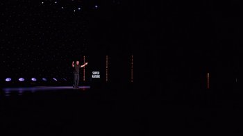 Movie still from “Ricky Gervais: SuperNature” (2022), directed by John L. Spencer – A man standing on a stage in front of a microphone; Extreme Wide shot, High angle