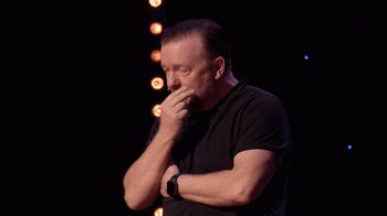 Movie still from “Ricky Gervais: SuperNature” (2022), directed by John L. Spencer – A man standing up with his hand to his face; Close Up shot, Over the shoulder angle