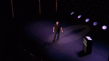 Movie still from “Ricky Gervais: SuperNature” (2022), directed by John L. Spencer – A man standing on a stage in front of an audience; Extreme Wide shot, Overhead angle